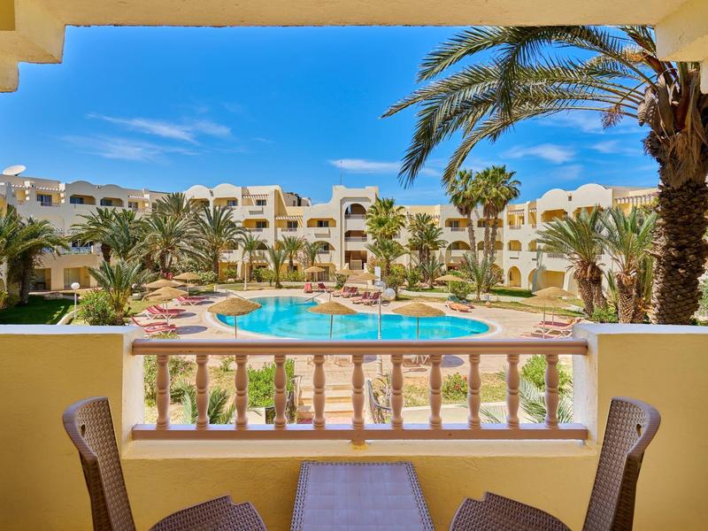 Vista desde un balcón hacia una piscina de hotel con palmeras y tumbonas bajo un cielo azul.