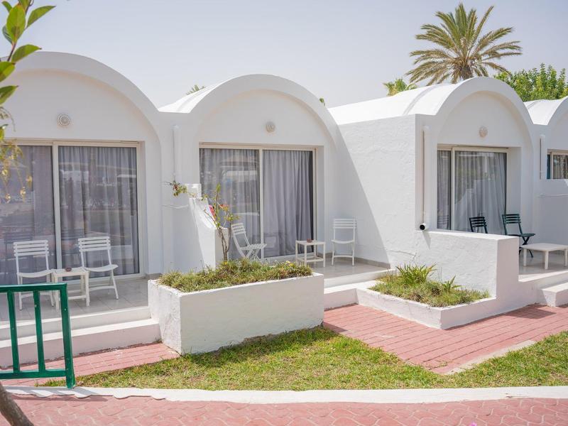 Bungalows blancos con pequeños jardines frontales, palmeras al fondo y cielo despejado.