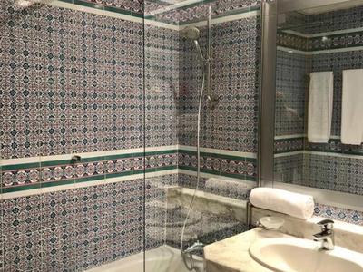 Nowoczesna łazienka z wanną, szklaną zasłoną prysznicową, białą umywalką i wzorzystymi kafelkami ściennymi.