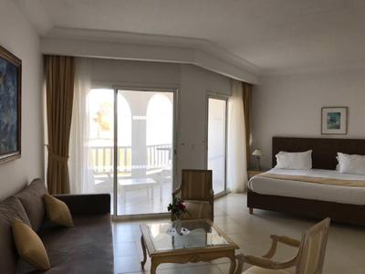 Jasny pokój hotelowy z podwójnym łóżkiem, sofą, fotelem i dostępem do balkonu z żółtymi poręczami.