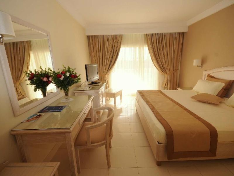 Jasny, przytulny pokój hotelowy z podwójnym łóżkiem, biurkiem i bukietem kwiatów na stole.