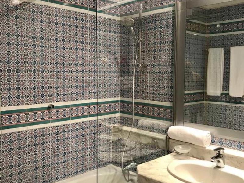 Nowoczesna łazienka z wanną, szklaną zasłoną prysznicową, białą umywalką i wzorzystymi kafelkami ściennymi.