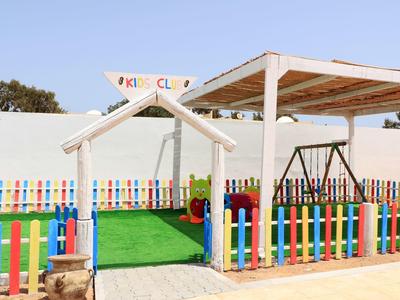 Bunter Kinderspielplatz mit Schaukeln, grünem Kunstrasen und buntem Lattenzaun unter blauem Himmel.