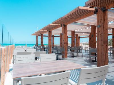 Strandrestaurant mit Holzbalken, weißen Tischen und Stühlen direkt am Meer unter klarem Himmel.