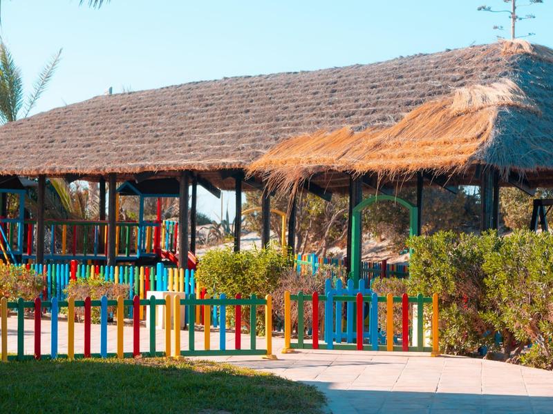 Colorful wooden fence and thatched roof pavilions in a garden area under a clear sky.