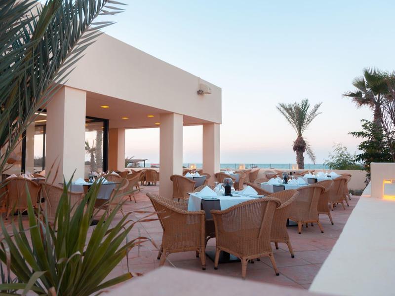 Hotel terrace with tables, chairs, palm trees, and a sunset view.
