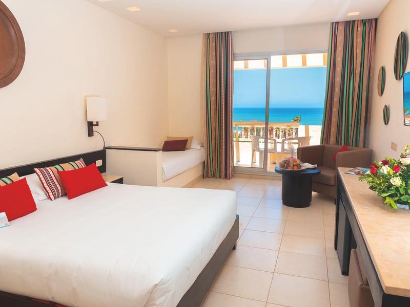 Bright hotel room with double bed, seating area, and balcony overlooking the sea.