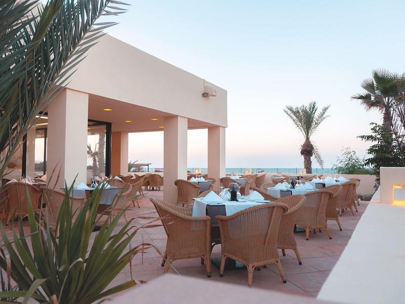 Terrace with modern chairs and tables beside a building, surrounded by palm trees at sunset.