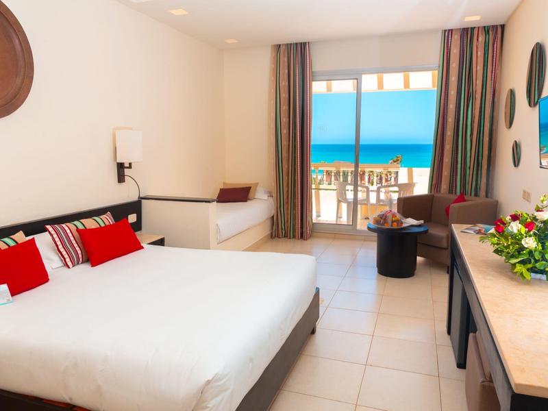 Bright hotel room with large bed, seating area, and balcony overlooking the sea.