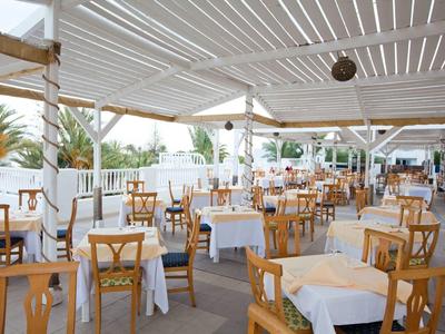 Helles, offenes Restaurant mit Holzmöbeln und weißen Tischen unter einem überdachten Außenbereich.