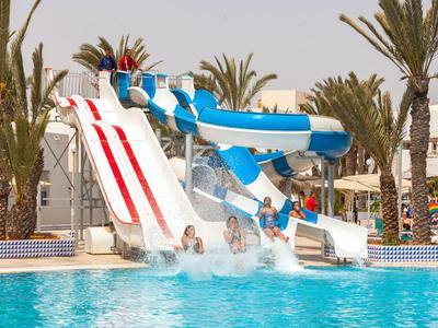 Kinder rutschen auf bunten Wasserrutschen in einem Pool mit Palmen im Hintergrund.