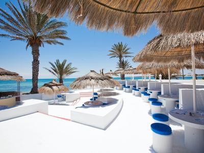 Strandbar mit weißen Sitzgelegenheiten, blauen Hockern und Palmen unter sonnigem Himmel am Meer.