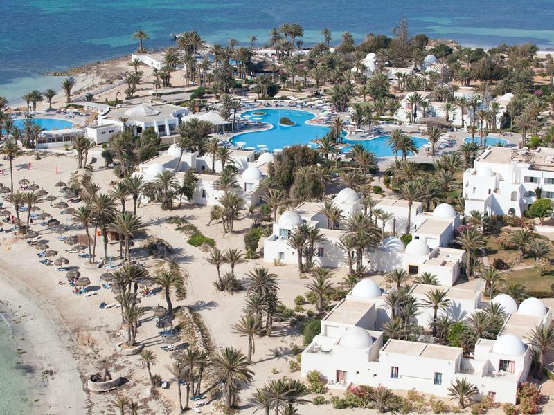 Luftaufnahme eines tropischen Resorts mit weißen Gebäuden, Pools und Palmen am Strand.