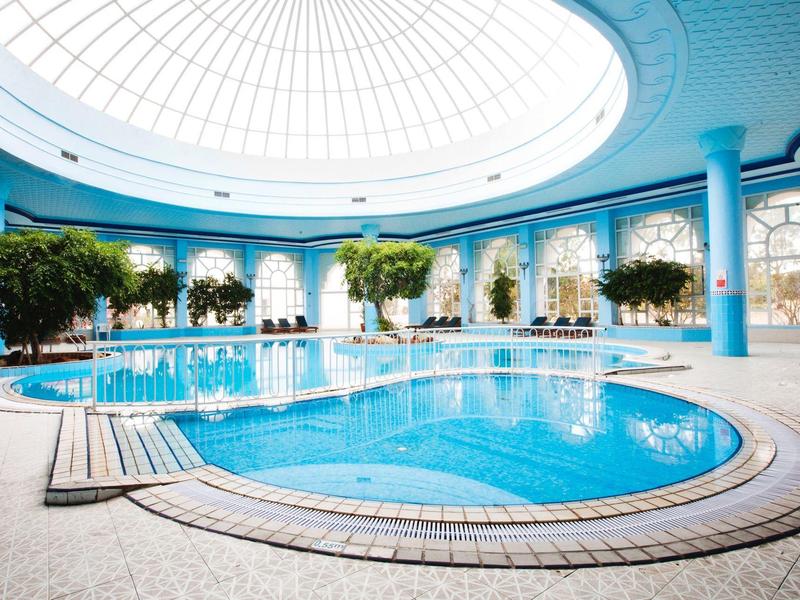 Großes Hallenbad mit blauem Wasser, Bäumen und großen Fenstern unter einer Kuppel