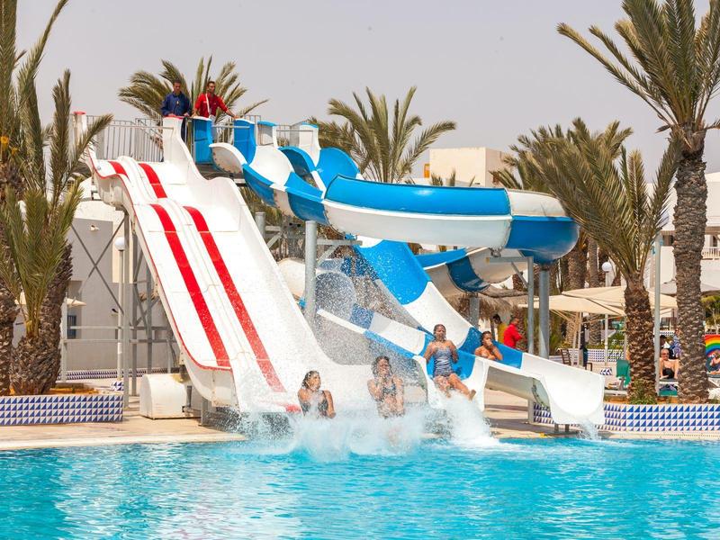 Kinder rutschen auf bunten Wasserrutschen in einem Pool mit Palmen im Hintergrund.