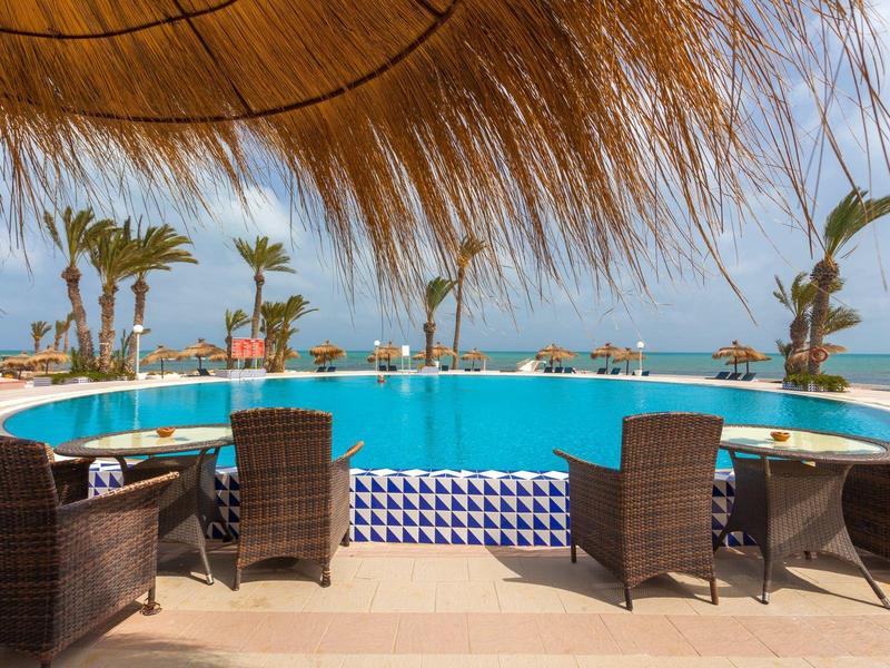 Blick auf einen Pool mit Sitzbereich unter einem Sonnendach und Palmen am Strand