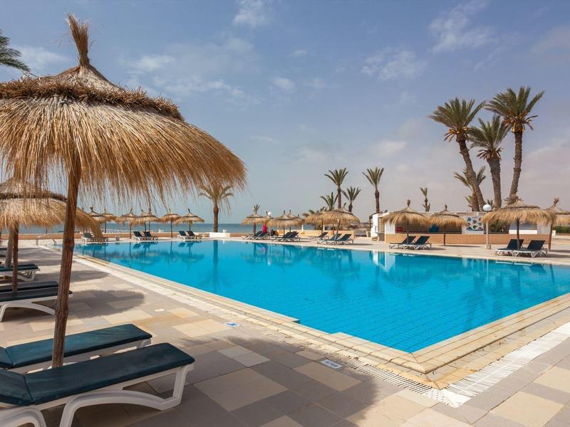 Großer Außenpool mit Liegestühlen und Strohmachschirmen unter blauem Himmel.