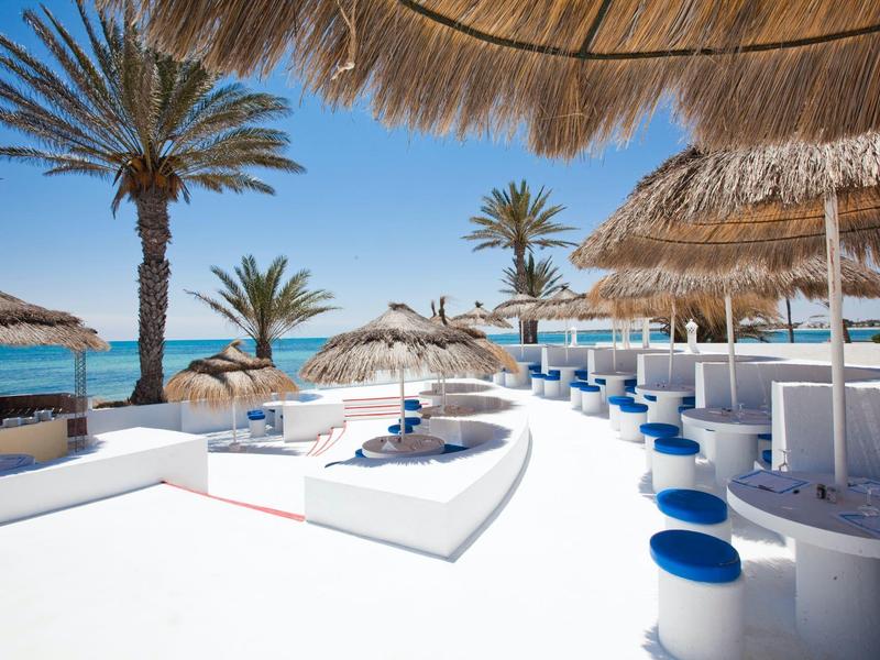 Strandbar mit weißen Sitzgelegenheiten, blauen Hockern und Palmen unter sonnigem Himmel am Meer.