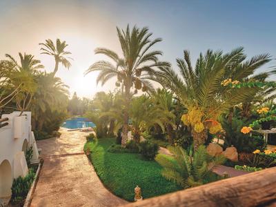 Sonniger Gartenweg mit Palmen und Pool neben einem Hotelgebäude.