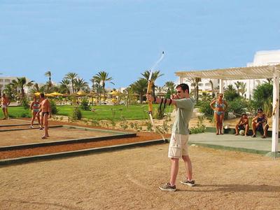 Menschen spielen Boule auf Sandplatz, Hintergrund mit Grünfläche und Palmen unter blauem Himmel.