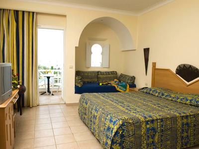 Hotelzimmer mit Doppelbett, Sofa, Fernseher, Balkon und gelb-blauen Vorhängen in hellen Farben.