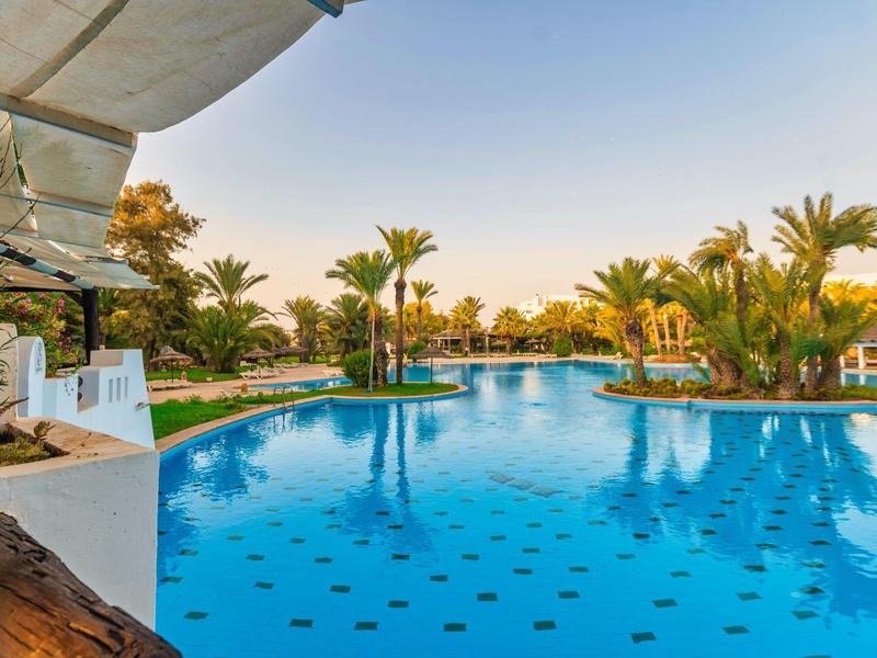 Großer, blauer Pool mit Palmen und grünen Pflanzen, klarer Himmel, entspannte Urlaubsatmosphäre.