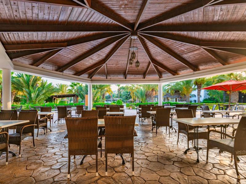 Überdachte Terrasse mit Holzdach, braunen Stühlen und Tischen auf Steinboden, grüne Umgebung.