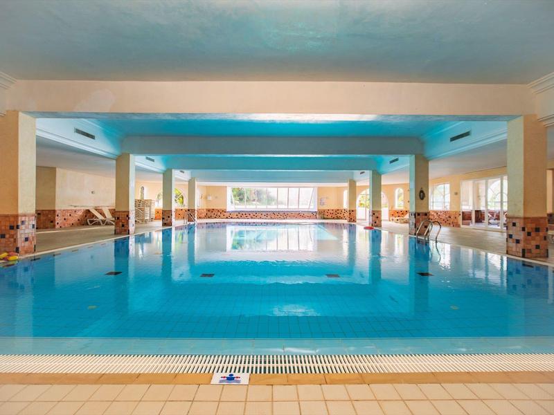 Großes, helles Hallenbad mit blauen Wasser und seitlichen Fenstern in einem Hotel.