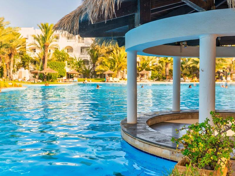 Großer Pool mit Rundbar, umgeben von Palmen und weißen Gebäuden unter blauem Himmel.