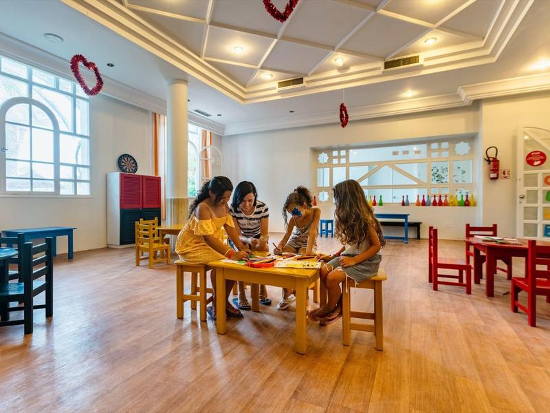 Kinder sitzen an einem Tisch in einem hellen, bunten Spielzimmer mit Holzfußboden.