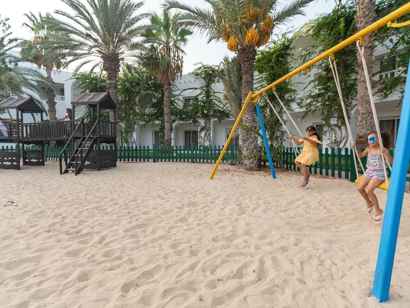 Spielplatz mit Sand, zwei Kindern auf Schaukeln, Palmen und Gebäuden im Hintergrund.