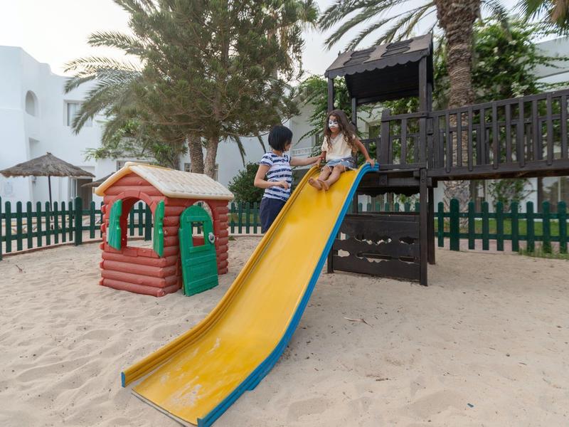 Kinder spielen auf einer gelben Rutsche im Sandkasten neben einem kleinen Spielhaus und Palmen.