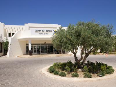 Eingang eines Hotels mit einem runden Blumenbeet und einem Baum davor unter klarem Himmel.