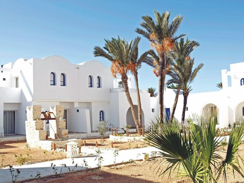 Witte gebouwen met blauwe deuren en palmbomen onder een heldere lucht in een hotelgebied.