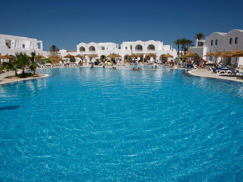 Groot blauw zwembad voor wit hotelgebouw bij heldere lucht.