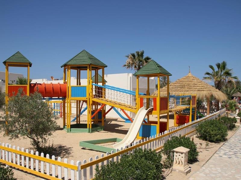 Kleurrijke speeltuin met glijbanen en klimnetten naast een hotel tuin bij een heldere lucht.