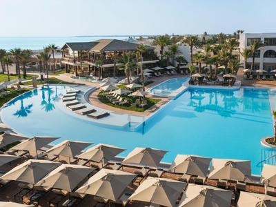 Großer Hotelpool mit Liegen, Palmen und Blick auf das Meer bei sonnigem Wetter.