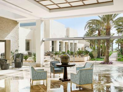 Bright hotel lobby with armchairs, tables, large windows, and view of palm trees outside.