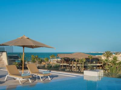 Área de piscina con tumbonas y sombrilla frente a vistas al mar y palmeras bajo cielo despejado