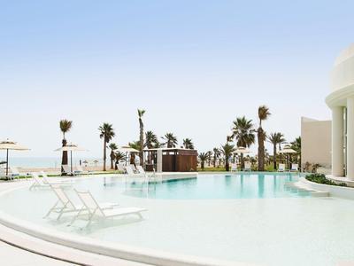 Heller Pool mit Liegestühlen und Palmen vor klarem Himmel und Meerblick.