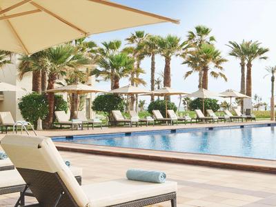 Hotelpool mit Liegestühlen und Sonnenschirmen, umgeben von Palmen an sonnigem Tag.