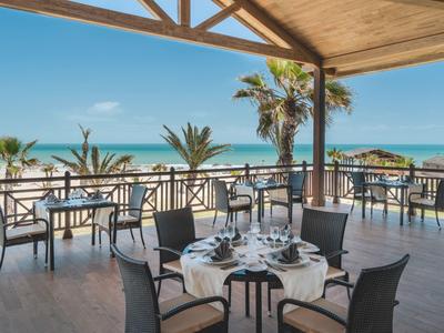 Terraza abierta con mesas y sillas dispuestas frente a palmeras y vista al mar.