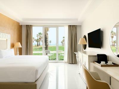 Helles Hotelzimmer mit Doppelbett, großem Fenster und Blick auf Palmen und Meer.