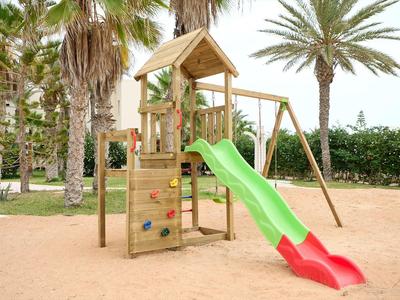 Área de juegos infantiles con tobogán, muro de escalar y columpio en zona arenosa con palmeras.