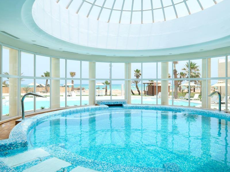 Piscina cubierta con techo de vidrio y vista al exterior playa y palmeras.