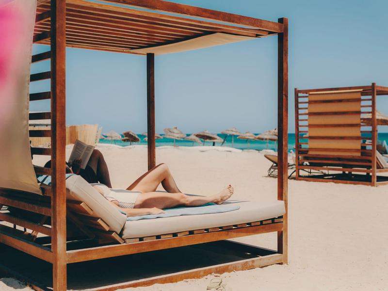 Persona relajándose en una cama de playa de madera bajo cielo azul en la playa.