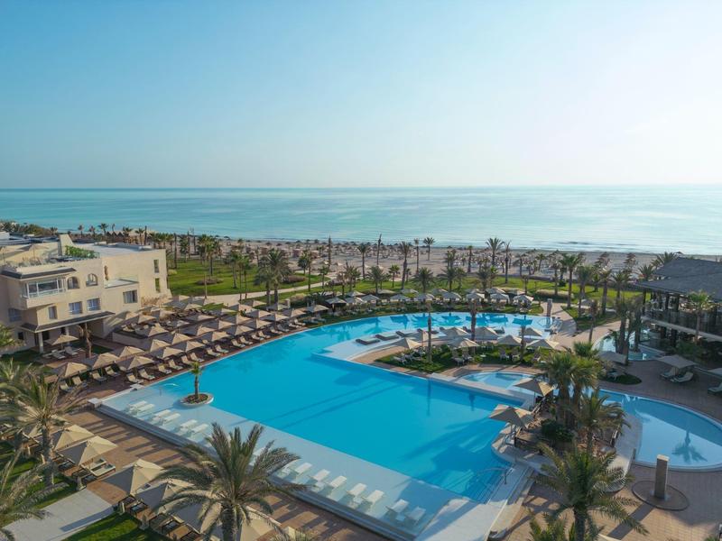Gran piscina de hotel con sombrillas y palmeras junto al mar bajo un cielo despejado.