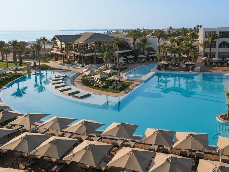 Gran piscina del hotel con tumbonas y palmeras bajo cielo despejado y vista al mar.