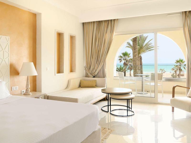 Habitación de hotel luminosa con cama doble, zona de estar y balcón con vista al mar y palmeras.