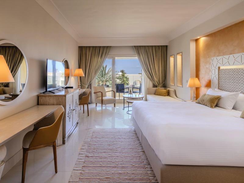 Habitación de hotel luminosa con cama grande, escritorio, televisión y balcón con vista al mar.